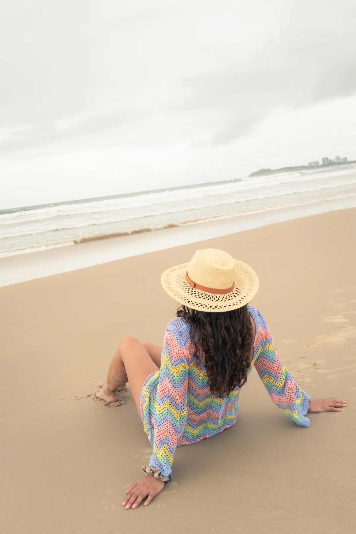 Mujer sentada de espaldas en playa con sombrero y tejido de colores