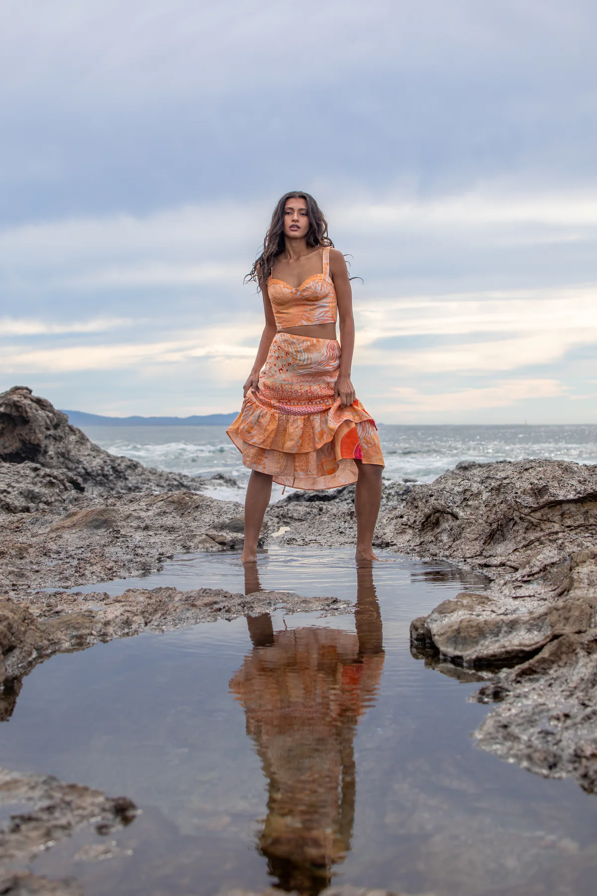 Retrato editorial con vestido naranja reflejado en una poza de roca